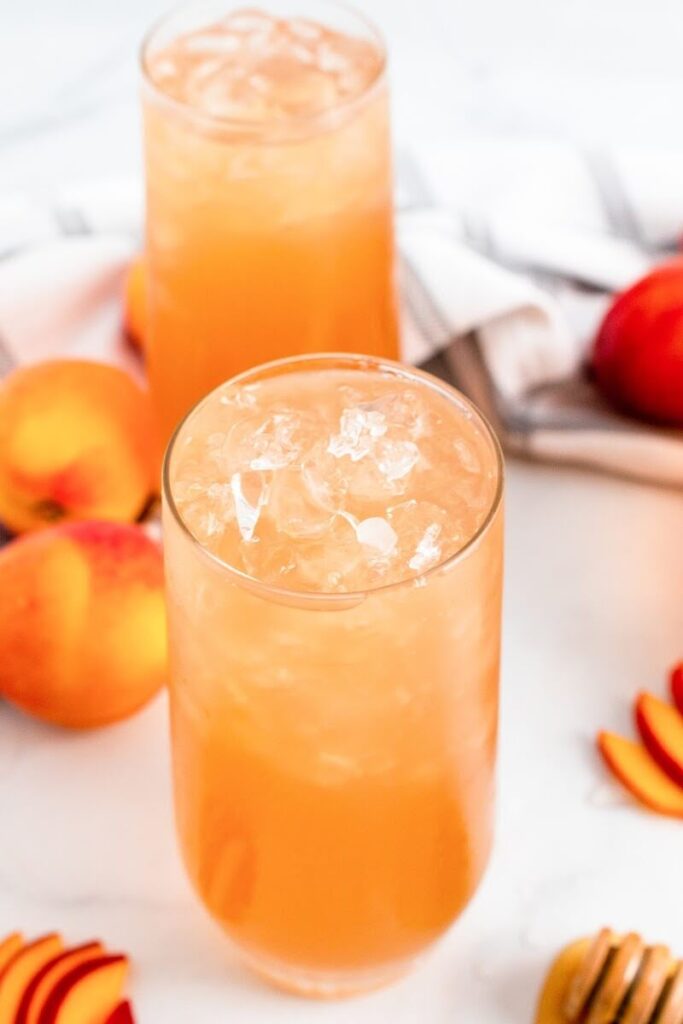 Two glasses full of iced peach tea sitting next to sliced peaches, fresh whole peaches and a honey dipper.