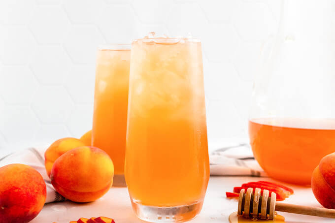 Two glasses full of iced peach tea sitting next to sliced peaches, fresh whole peaches, a honey dipper and a pitcher half full of peach tea.