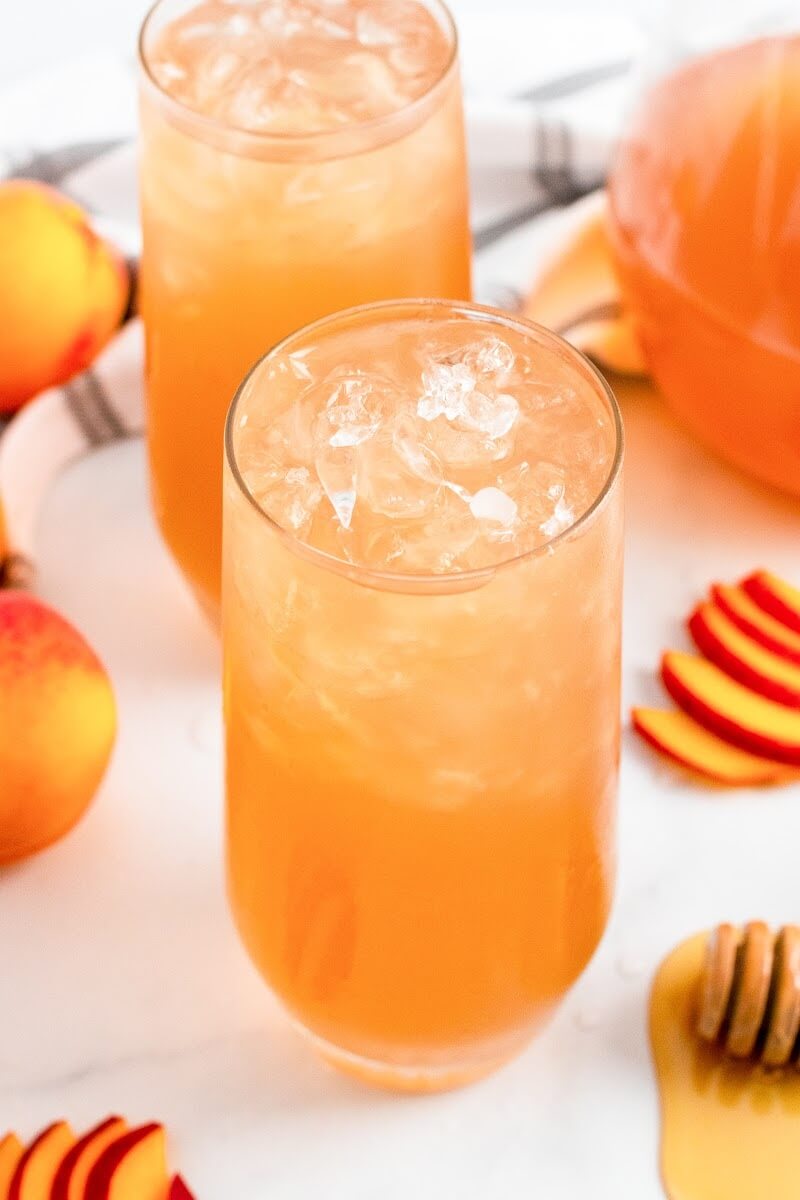 Two glasses full of iced peach tea sitting next to sliced peaches, fresh whole peaches, a honey dipper and a pitcher half full of peach tea.