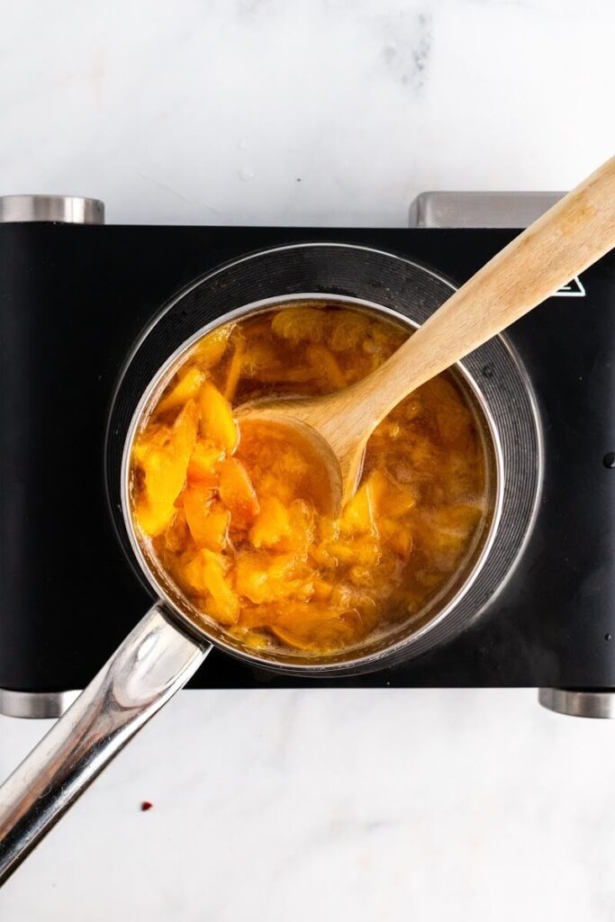A saucepan with cooked peaches and a wooden spoon stirring it sitting on top of an electric stovetop.