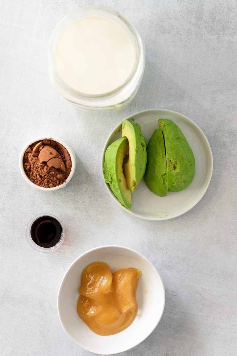 Overhead shot o quartered and peeled avocados on a small plate, jar of milk, small bowl of cocoa powder, small bowl of honey and tiny bowl of vanilla extract.