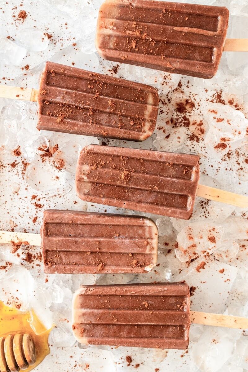 5 fudgesicle popsicles dusted with cocoa powder sitting on top of ice, next to a honey dipper with honey on it.
