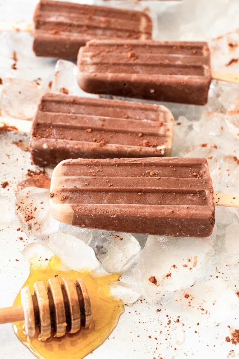 4 fudgesicle popsicles dusted with cocoa powder sitting on top of ice, next to a honey dipper with honey on it.