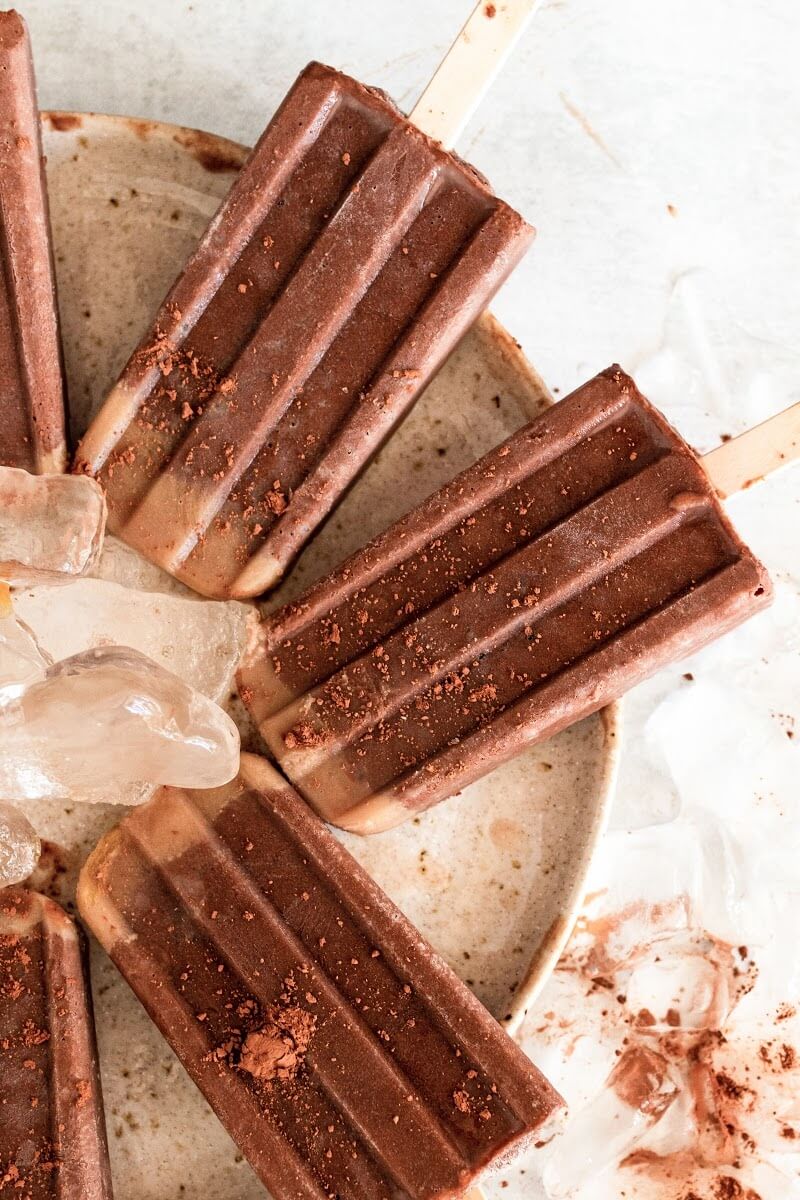 Fudgesicle popsicles dusted with cocoa powder sitting on a plate with ice.
