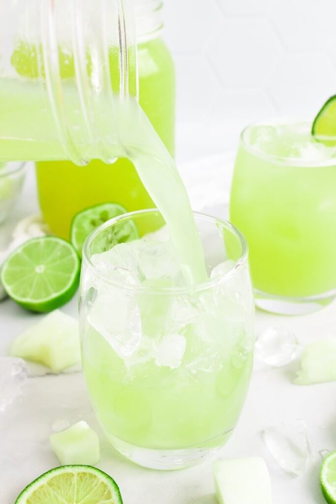 Green-colored drink being poured into a glass filled with ice, next to another glass full of the iced green drink with a slice of fresh lime on the rim, surrounded by chunks of honeydew melon, ice cubes, fresh lime slices and a jar filled with the drink.