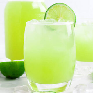 Glass filled with iced green-colored drink with a slice of fresh lime on the rim, surrounded by chunks of honeydew melon, ice cubes, fresh lime slices, another glass filled with the iced green drink and a jar filled with the drink.