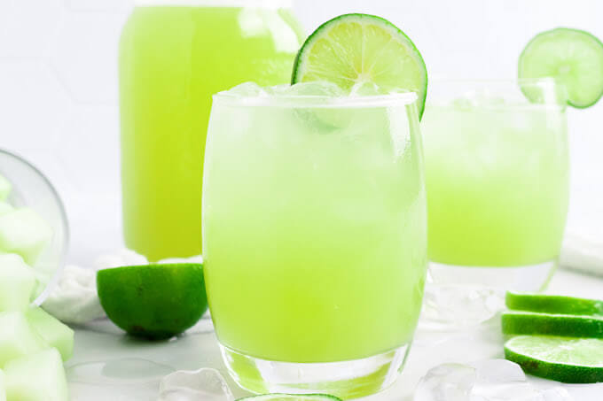 Glass filled with iced green-colored drink with a slice of fresh lime on the rim, surrounded by chunks of honeydew melon, ice cubes, fresh lime slices, another glass filled with the iced green drink and a jar filled with the drink.