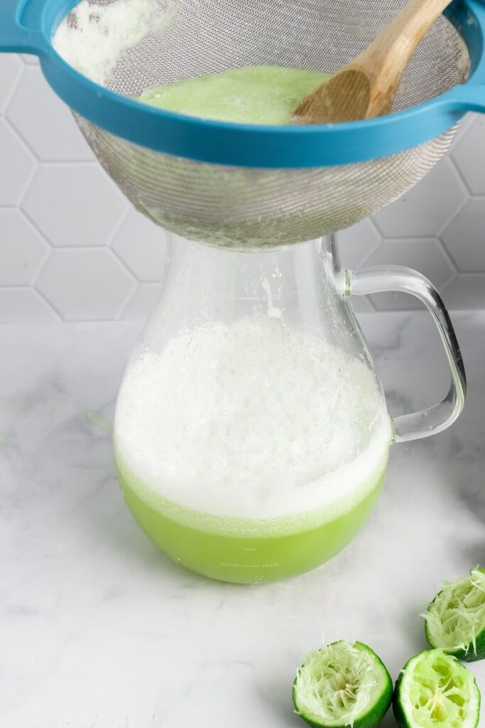 A glass pitcher half filled with a green colored drink, with a one mesh strainer sitting on top of the pitcher filled with juice pulp and 3 halves of juiced limes sitting on the counter next to the pitcher.