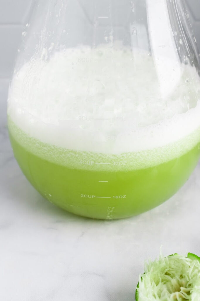 A closeup of a glass pitcher half filled with a green colored drink and a half juiced lime sitting on the counter next to the pitcher.