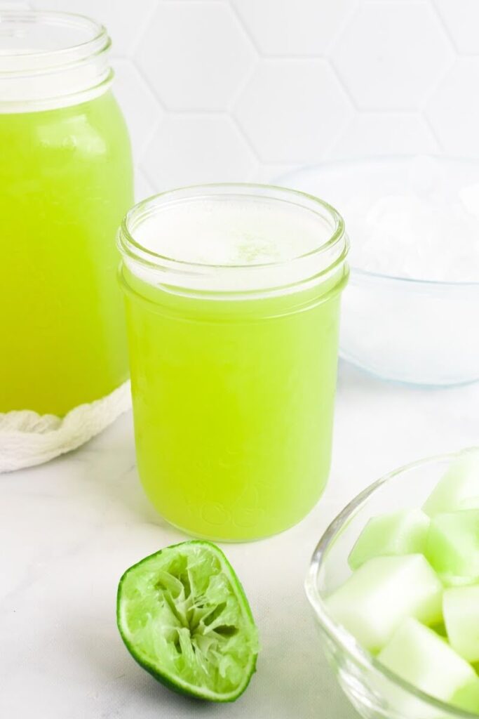 Two mason jars filled with a green-colored drink sitting next to a bowl filled with ice, a bowl with chunks of honeydew melon and a half of a juiced lime.