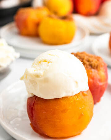 Cooked stuffed peaches sitting on plates, one topped with a scoop of vanilla ice cream, sitting next to fresh peaches, an ice cream scooper full of ice cream and an Instant Pot.