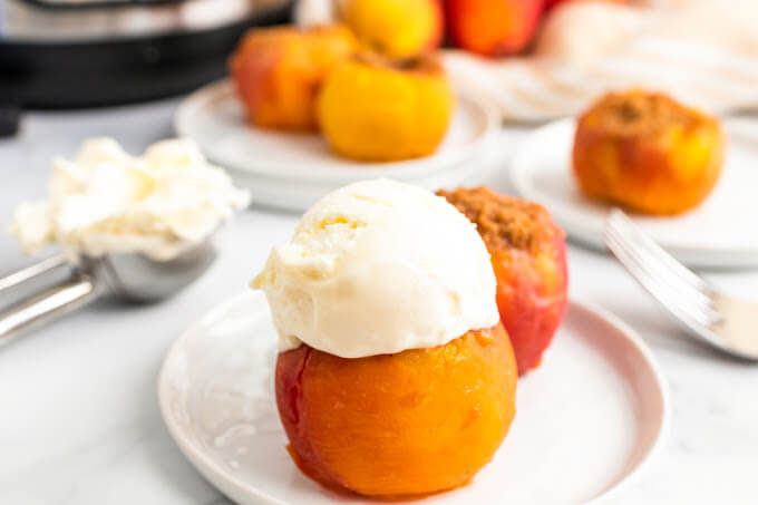 Cooked stuffed peaches sitting on plates, one topped with a scoop of vanilla ice cream, sitting next to fresh peaches, an ice cream scooper full of ice cream and an Instant Pot.