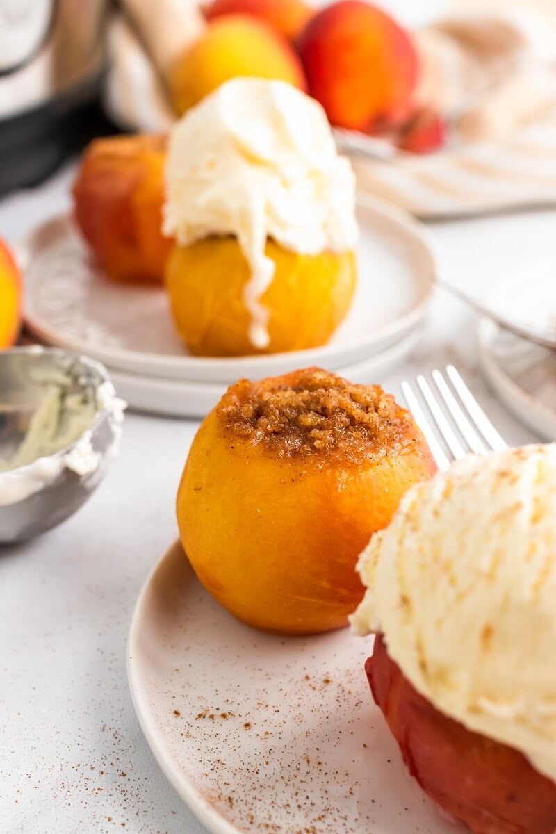 Cooked stuffed peaches sitting on plates, two topped with a scoop of vanilla ice cream, sitting next to fresh peaches, an ice cream scooper and an Instant Pot.