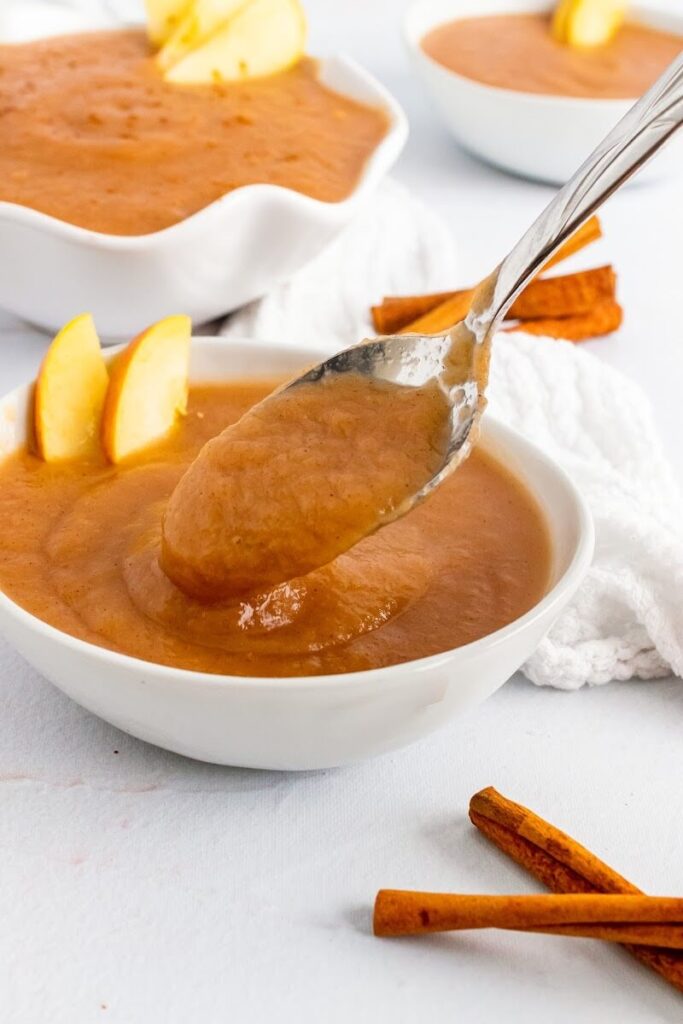 A close up of a spoon full of homemade applesauce, with three bowls filled with homemade applesauce with slices of apple, sitting next to a towel and cinnamon sticks in the background.