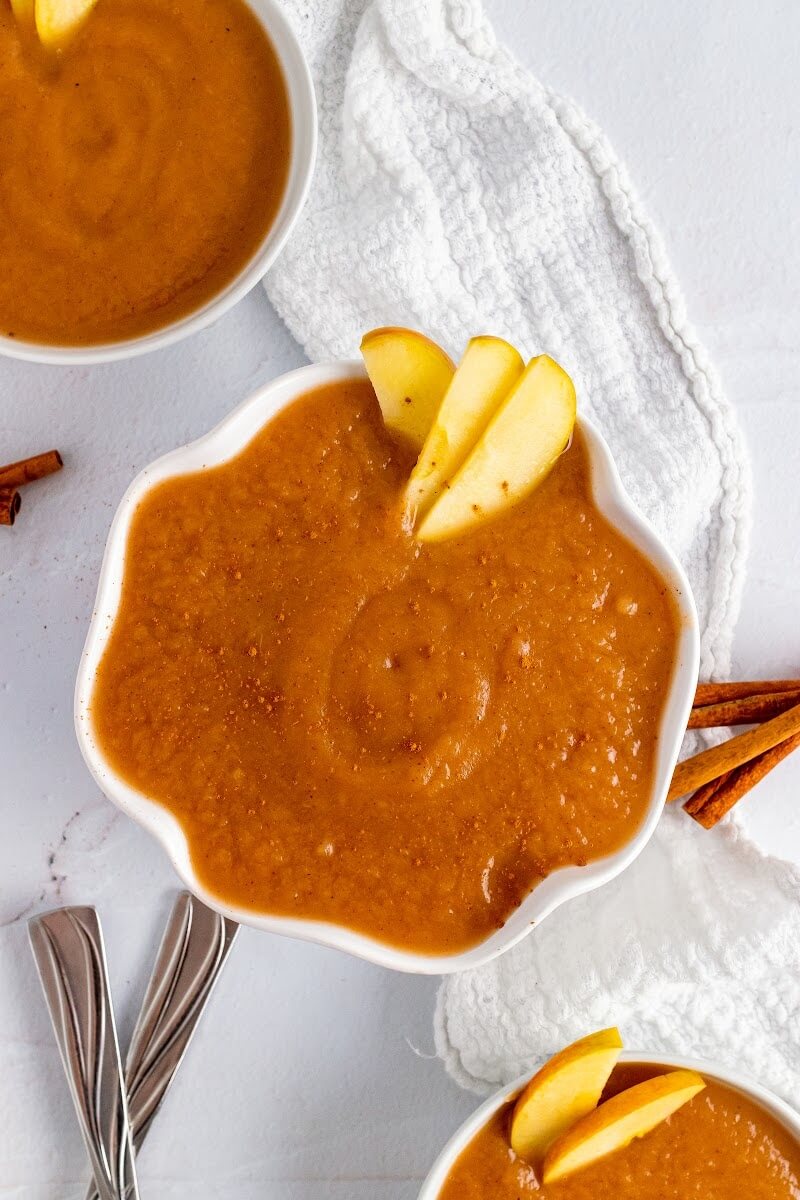 Three bowls filled with homemade applesauce with slices of apple, one dusted with ground cinnamon, sitting next to a towel, spoons and cinnamon sticks.