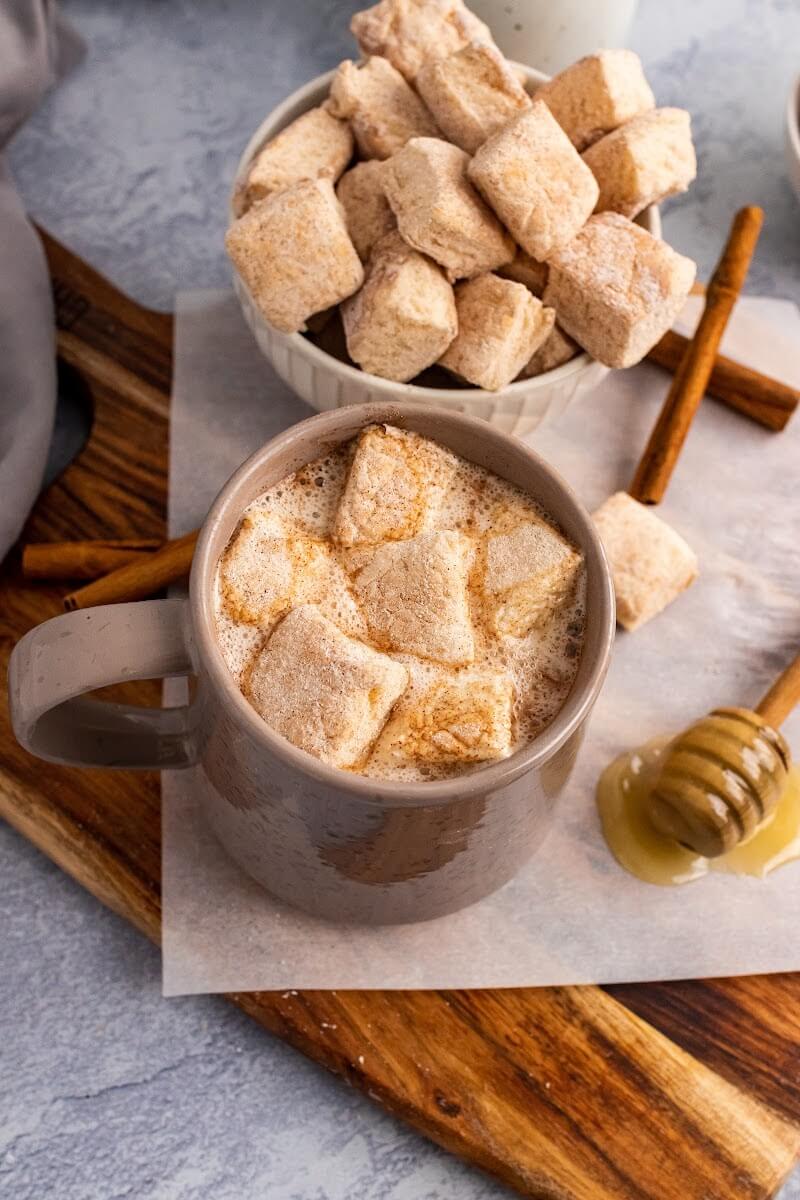 A mug full of hot chocolate topped with homemade marshmallows, sitting next to two bowls full of homemade marshmallows, next to cinnamon sticks, a honey dipper with honey, and more homemade marshmallows sitting on a cutting board.