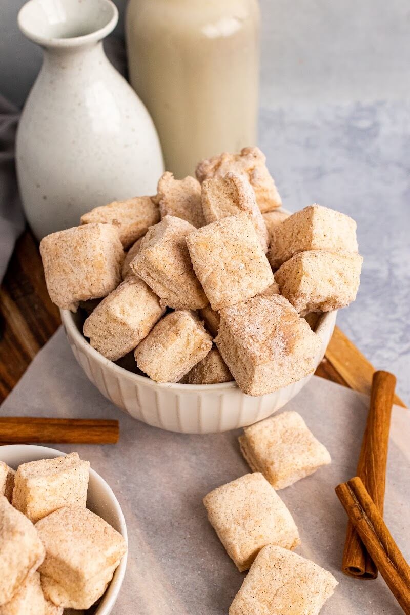 Two bowls full of homemade marshmallows, sitting next to cinnamon sticks, two small jars and more homemade marshmallows sitting on a cutting board.