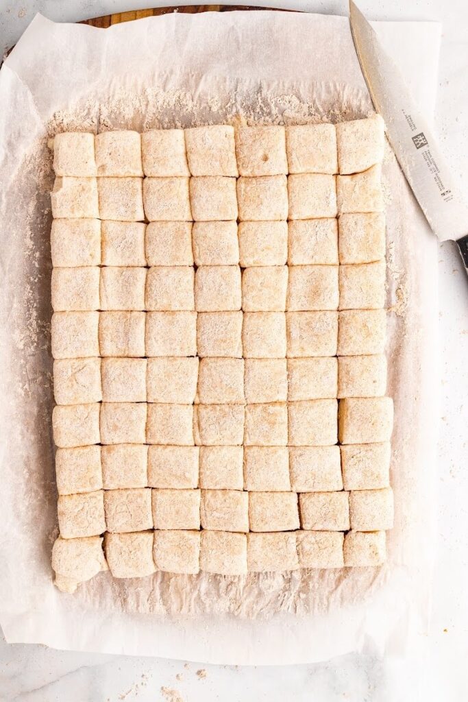 Parchment paper sitting on top of a cutting board and counter with a chef's knife and square-cut homemade marshmallows that are fully covered and dusted with an arrowroot-ground cinnamon mixture on top.