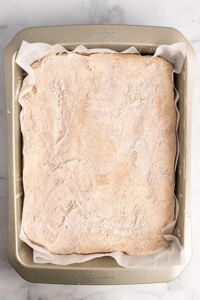 A rectangle baking pan lined with parchment paper and filled with uncut homemade marshmallows that are fully covered and dusted with an arrowroot-ground cinnamon mixture on top.