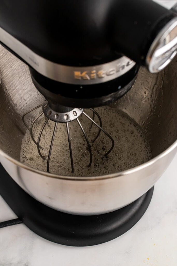 A standing mixer fitted with the whisk attachment and stainless steel mixing bowl, with a bubbly, frothy mixture inside the bowl.