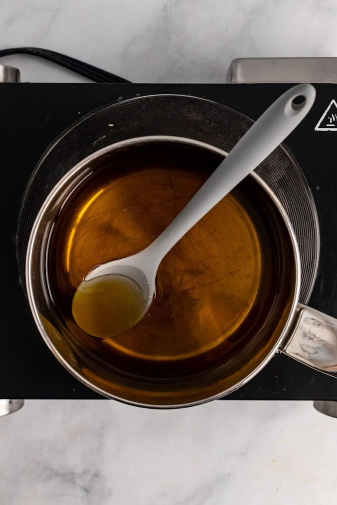 A small saucepan with a spoon, filled with honey and water, sitting on a stovetop burner.
