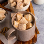 A mug full of hot chocolate topped with homemade marshmallows, sitting next to two bowls full of homemade marshmallows, next to cinnamon sticks, a honey dipper with honey, one small jar and more homemade marshmallows sitting on a cutting board.