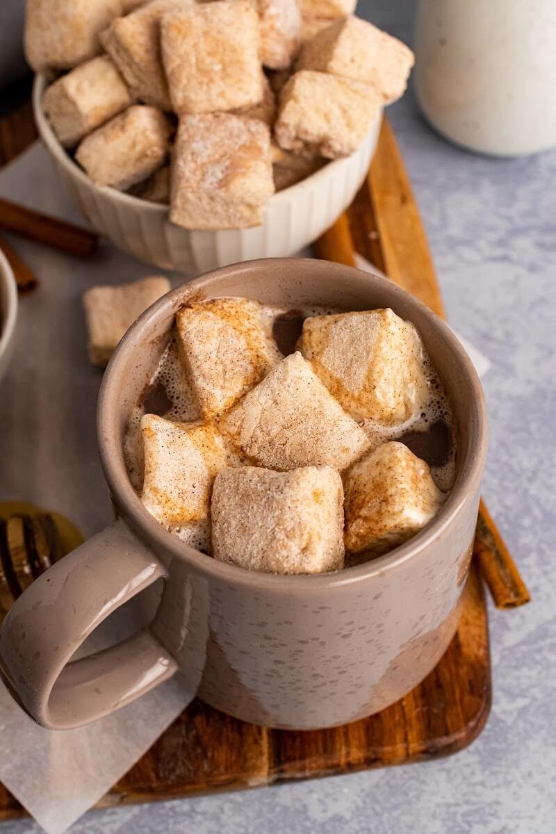 A mug full of hot chocolate topped with homemade marshmallows, sitting next to two bowls full of homemade marshmallows, next to cinnamon sticks, a honey dipper with honey, one small jar and more homemade marshmallows sitting on a cutting board.