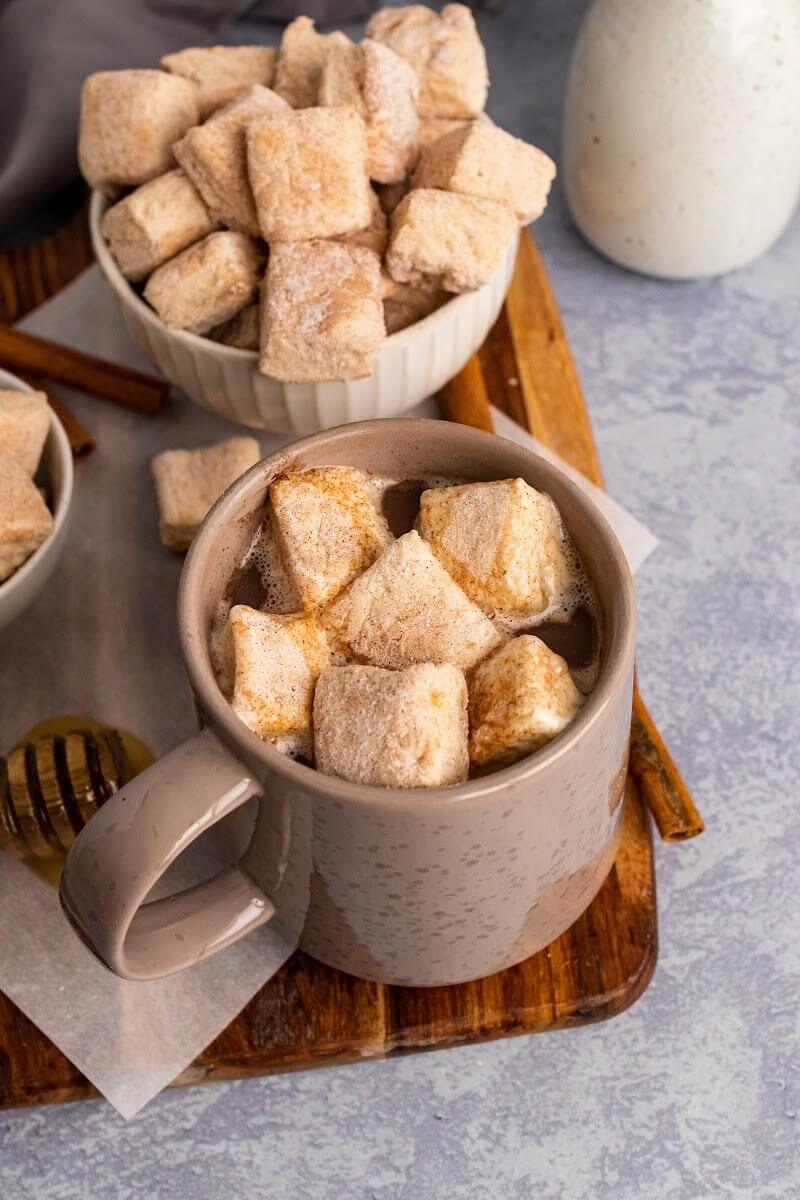 A mug full of hot chocolate topped with homemade marshmallows, sitting next to two bowls full of homemade marshmallows, next to cinnamon sticks, a honey dipper with honey, one small jar and more homemade marshmallows sitting on a cutting board.