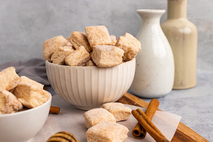 Two bowls full of homemade marshmallows, sitting next to cinnamon sticks, a honey dipper with honey, two small jars and more homemade marshmallows sitting on a cutting board.