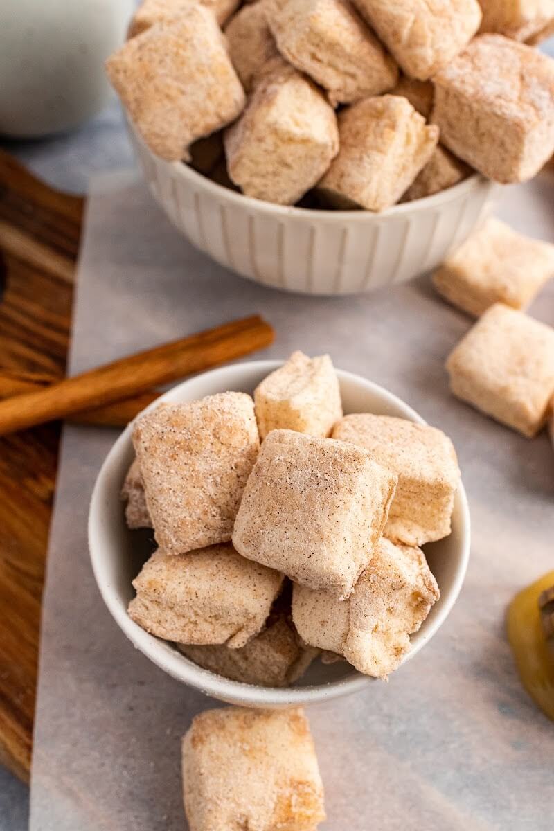 Two bowls full of homemade marshmallows, sitting next to cinnamon sticks, a honey dipper with honey, and more homemade marshmallows sitting on a cutting board.