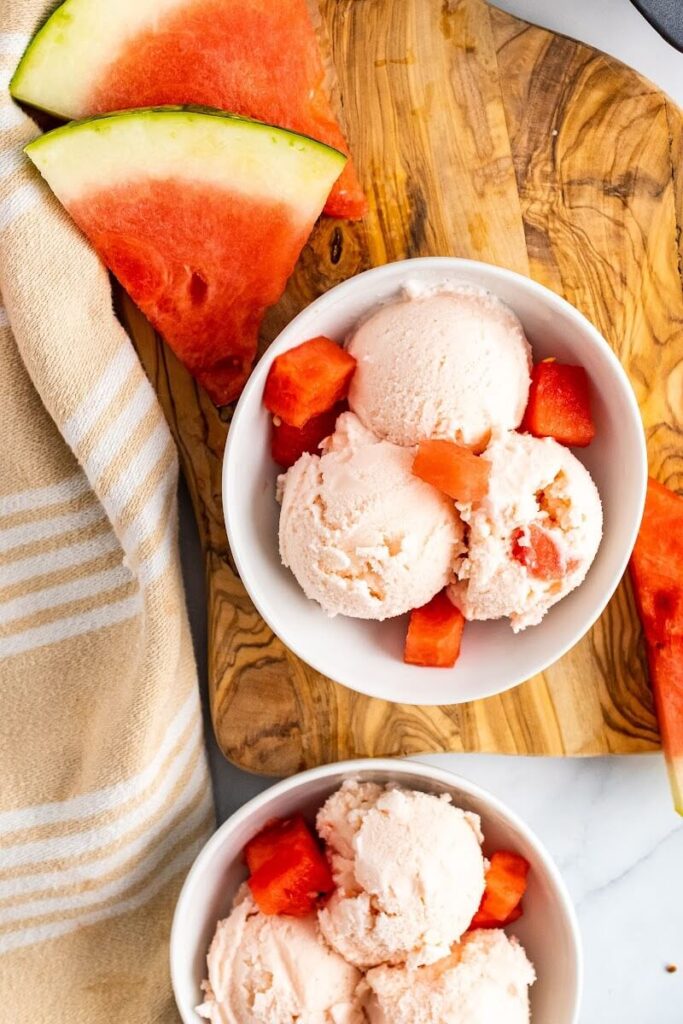 Two bowls with 3 scoops of watermelon ice cream and fresh watermelon chunks sitting on a cutting board next to freshly sliced watermelon and a cloth kitchen towel.