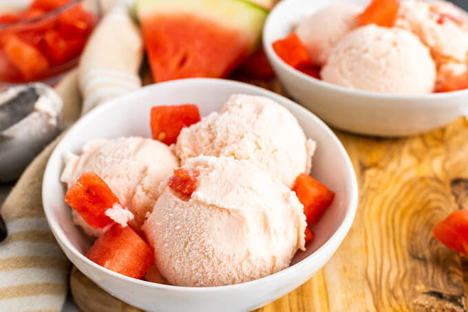Two bowls with 3 scoops of watermelon ice cream and fresh watermelon chunks sitting on a cutting board next to freshly sliced watermelon, a cloth kitchen towel and ice cream scooper.