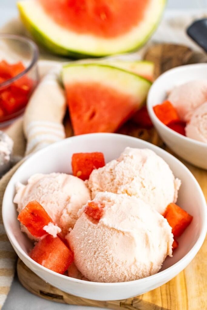 Bowl with 3 scoops of watermelon ice cream and fresh watermelon chunks sitting on a cutting board next to freshly sliced watermelon.