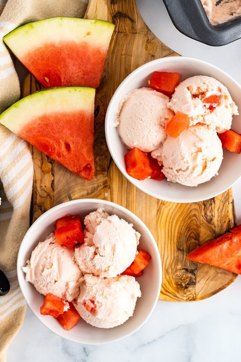Two bowls with 3 scoops of watermelon ice cream and fresh watermelon chunks sitting on a cutting board next to freshly sliced watermelon and a cloth kitchen towel.