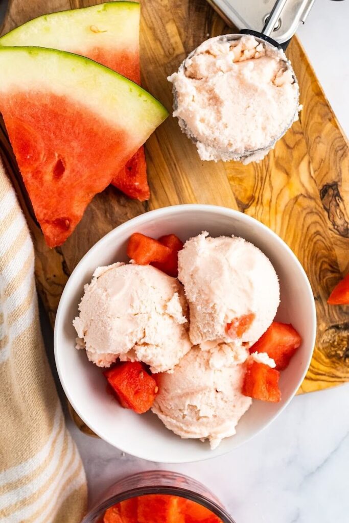Bowl with 3 scoops of watermelon ice cream and fresh watermelon chunks sitting on a cutting board next to freshly sliced watermelon, a cloth kitchen towel and a ice cream scooper full of ice cream.