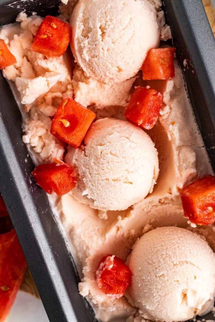 Loaf pan filled with homemade watermelon ice cream, 3 individual scoops of ice cream and fresh watermelon chunks.