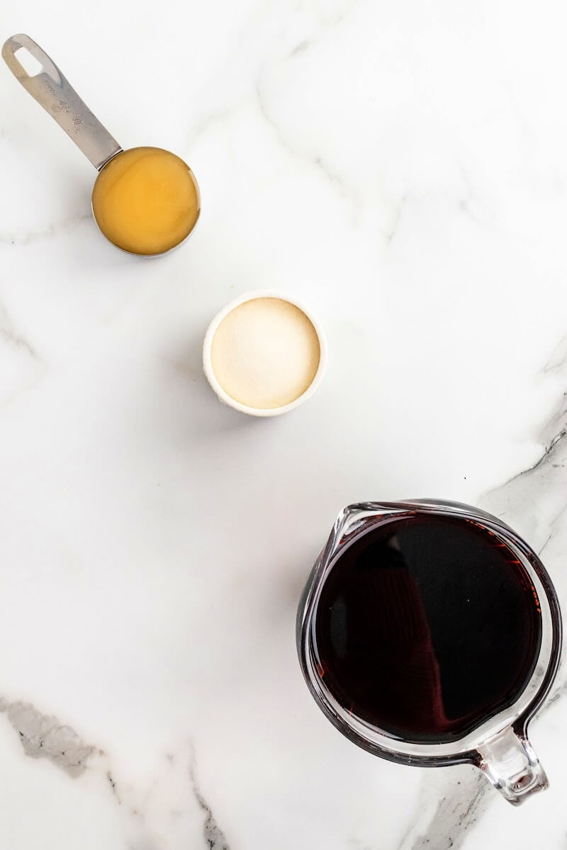 A overhead shot of a measuring cup filled with honey, a small bowl filled with gelatin and a glass measuring cup filled with dark purple juice.