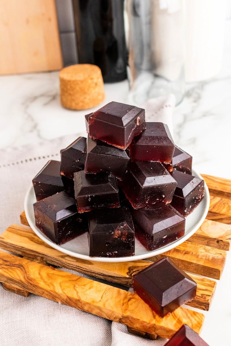 Plate with stacks of dark purple homemade gelatin gummies, sitting on top of a wooden board on top of a marble counter, with bottles in the background.