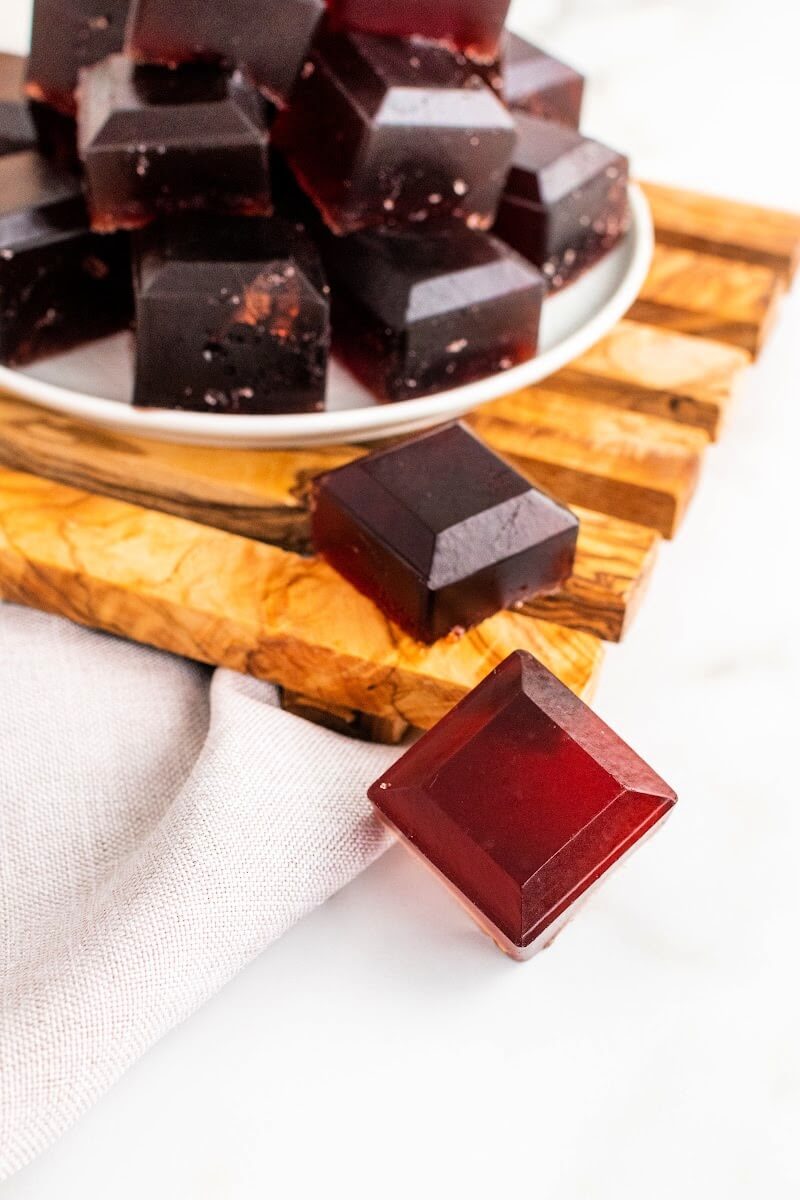 Plate with stacks of dark purple homemade gelatin gummies, sitting on top of a wooden board, with 1 gelatin gummy square sitting on the wood and 1 gelatin gummy sitting on the counter.