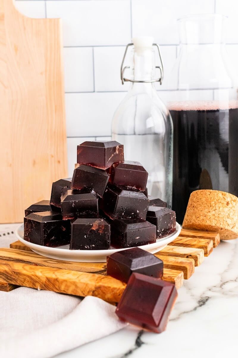 Plate with stacks of dark purple homemade gelatin gummies, sitting on top of a wooden board on top of a marble counter, with bottles in the background, one filled with dark juice, sitting next to a cutting board.