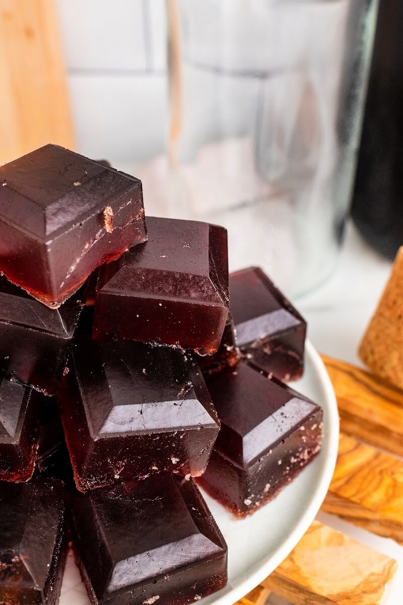 Plate with stacks of dark purple homemade gelatin gummies, sitting on top of a wooden board on top of a counter, with bottles in the background, one filled with dark juice.