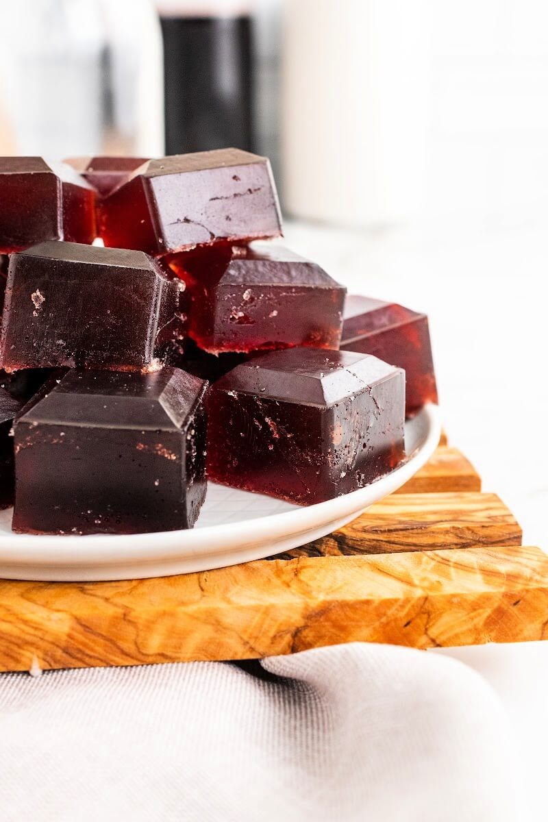 Plate with stacks of dark purple homemade gelatin gummies, sitting on top of a wooden board on top of a marble counter, with bottles in the background, one filled with dark juice.