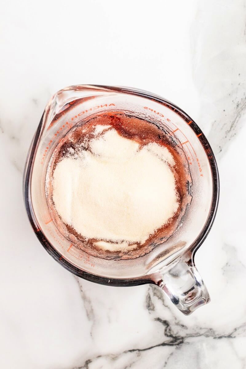 A glass measuring cup filled with a dark purple liquid, topped with gelatin, sitting on top of a marble counter.