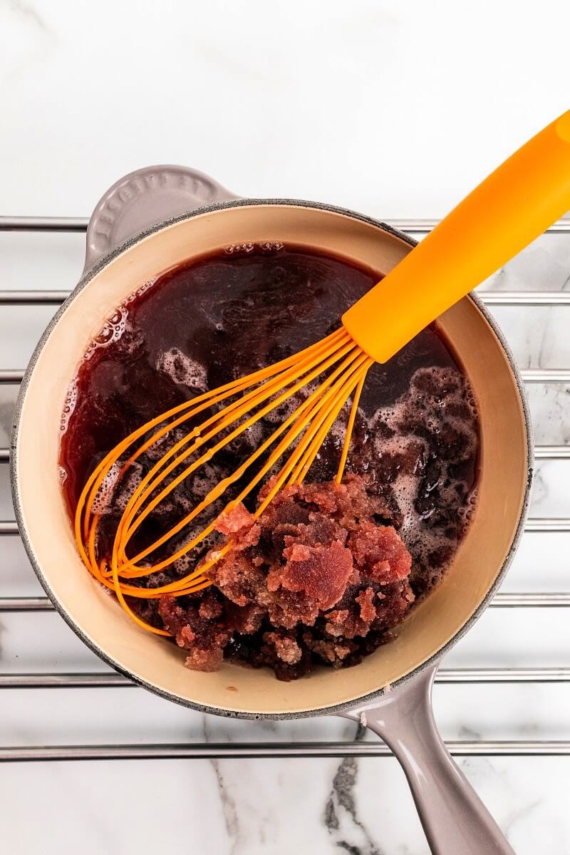 Saucepan sitting on a metal rack with a whisk stirring a dark purple liquid and clumps of bloomed gelatin.