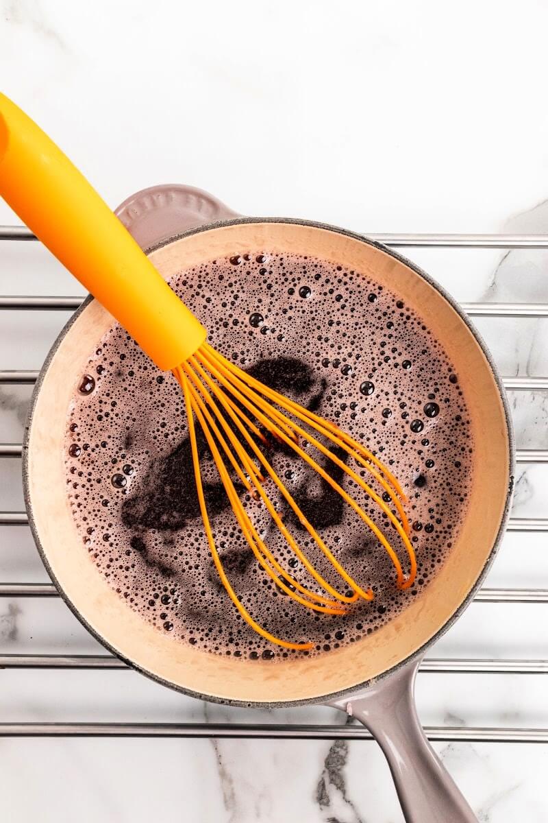 Saucepan sitting on a metal rack with a whisk stirring a frothy dark purple liquid.