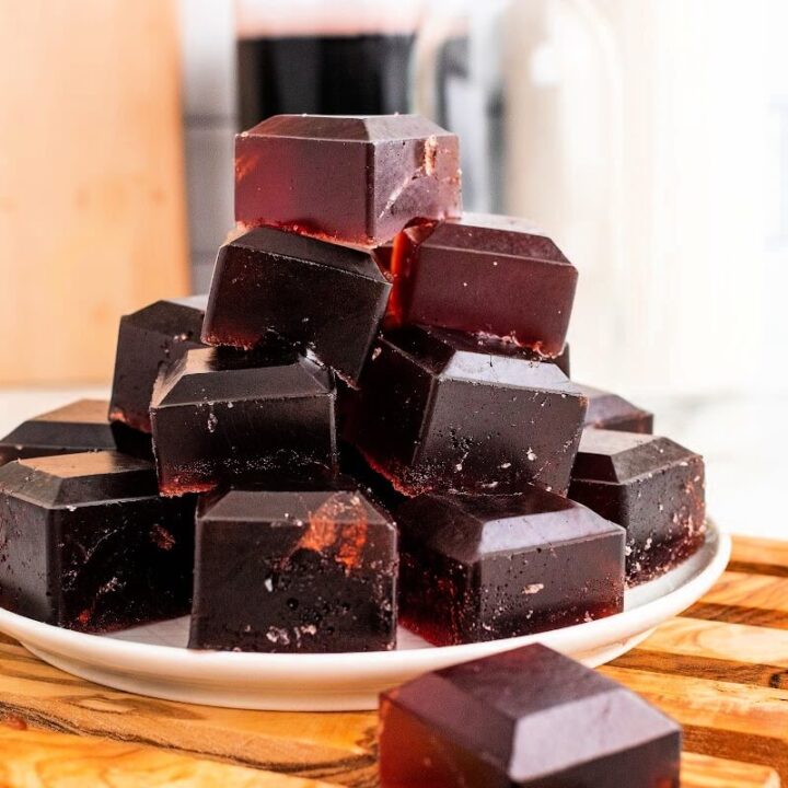 Plate with stacks of dark purple homemade gelatin gummies, sitting on top of a wooden board on top of a marble counter, with bottles in the background, one filled with dark juice, sitting next to a cutting board.