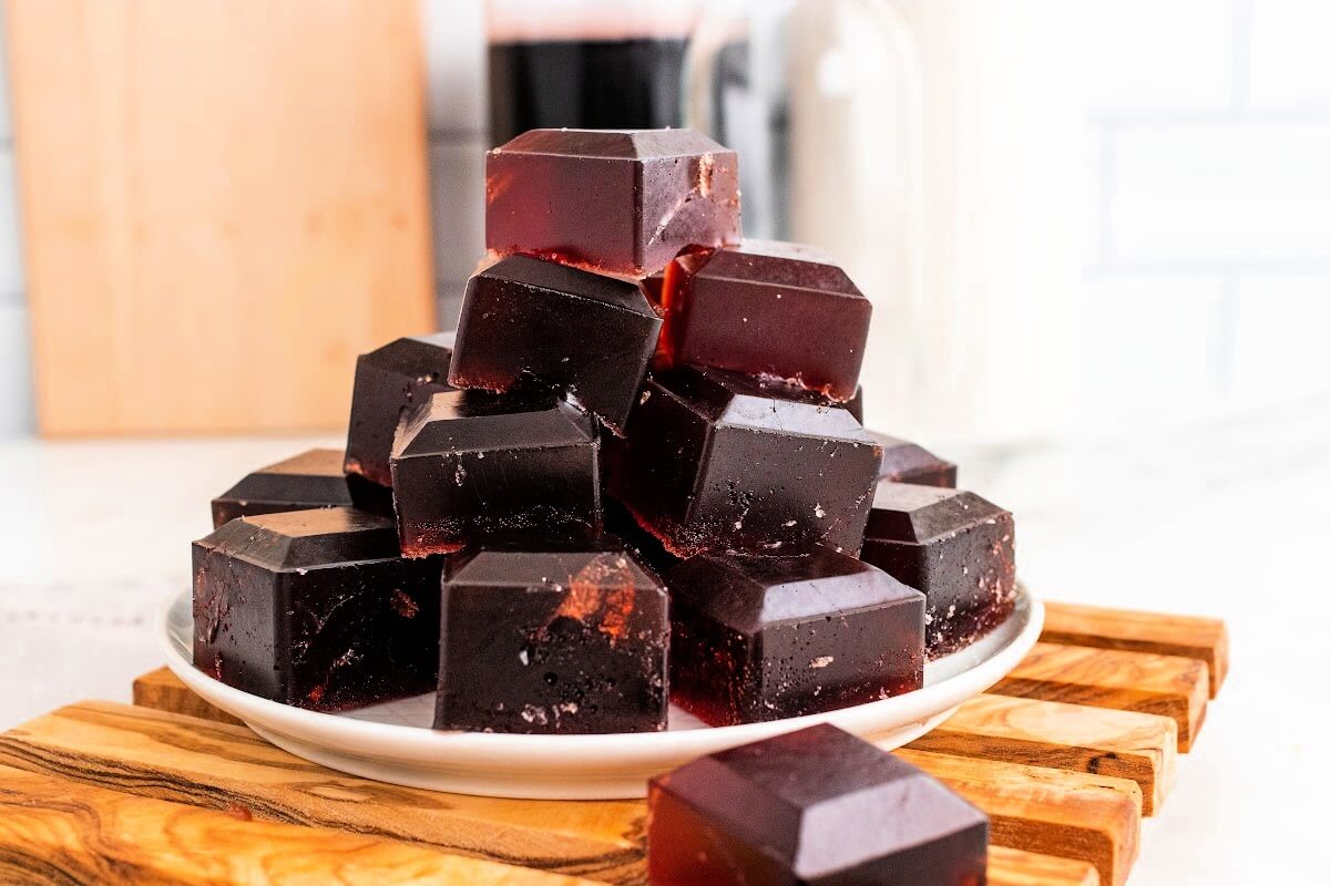 Plate with stacks of dark purple homemade gelatin gummies, sitting on top of a wooden board on top of a marble counter, with bottles in the background, one filled with dark juice, sitting next to a cutting board.