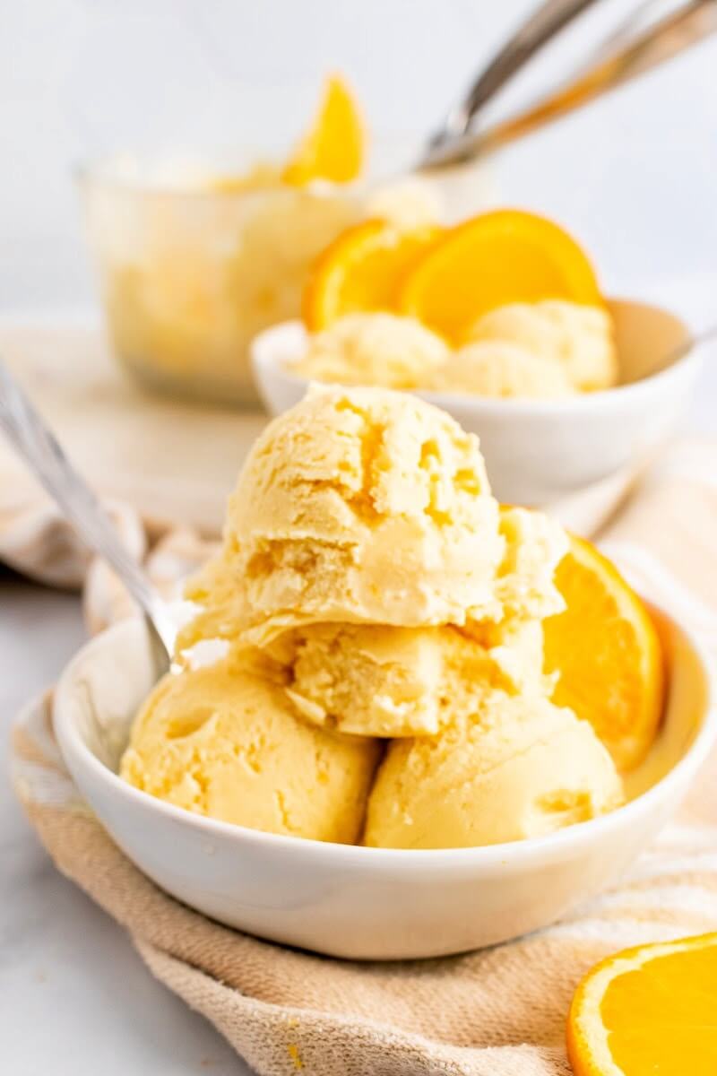 Two bowls with scoops of orange ice cream, a spoon and fresh orange slices sitting next to a large bowl full of ice cream and an ice cream scooper.