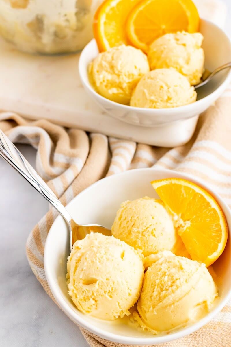 Two bowls with scoops of orange ice cream, a spoon and fresh orange slices sitting on top of a cloth kitchen towel.