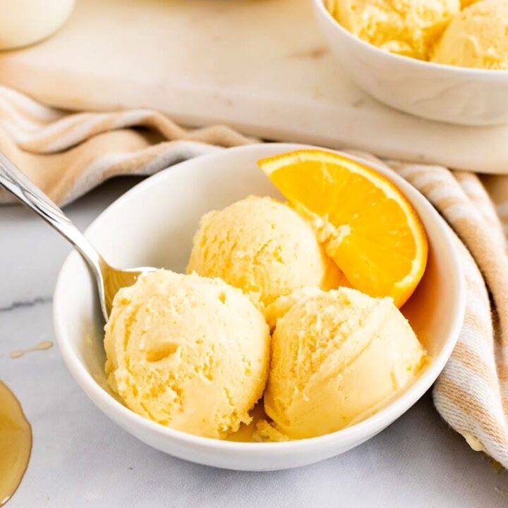 Bowl with scoops of orange ice cream, a spoon and fresh orange slices and honey spilled next to it.
